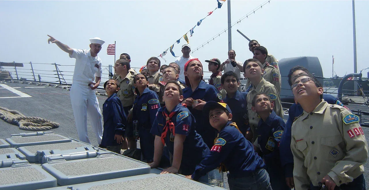Muslim scouts in uniform aboard a Navy ship, looking up as a sailor points to the sky